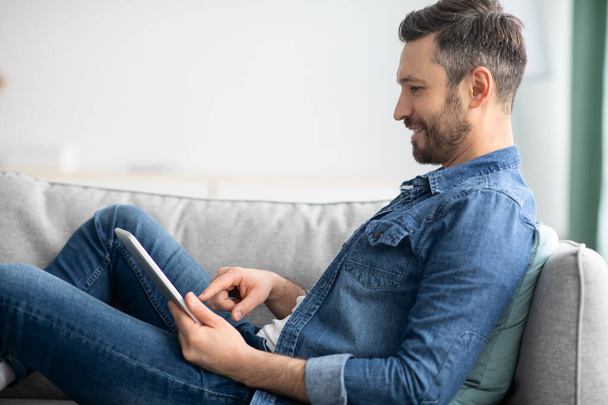 Щасливий чоловік сидить на дивані вдома, використовуючи цифровий планшет
 - Фото, зображення