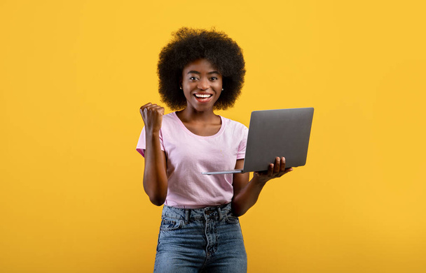 Euphorische schwarze Dame feiert Erfolg mit Laptop und reckt geballte Faust über gelbem Hintergrund - Foto, Bild