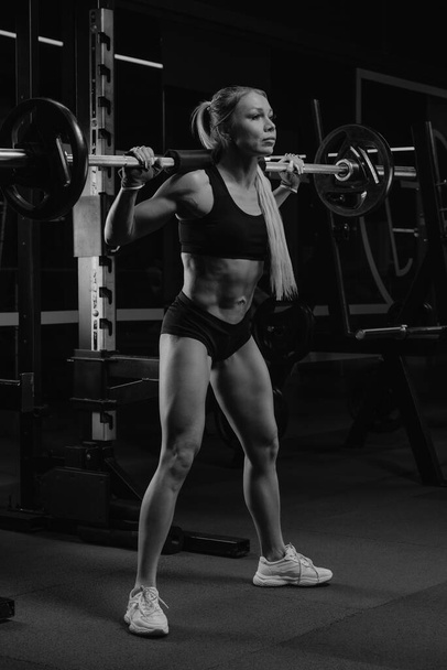 Une femme sportive aux cheveux blonds commence à squatter avec un haltère près du carré de squat dans une salle de gym. Une fille fait un entraînement de jambe. - Photo, image