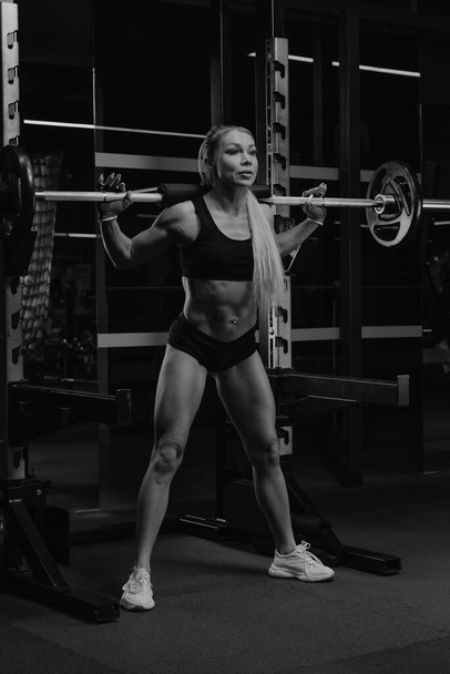 A sporty woman with blonde hair is squatting with a barbell near the squat rack in a gym. A girl is doing a leg workout. - Photo, Image