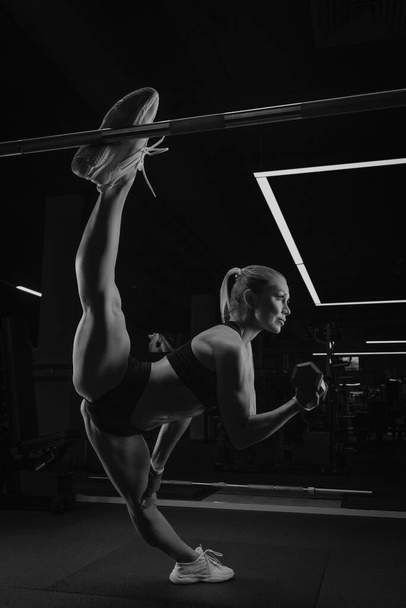 Eine sportliche Frau mit langen blonden Haaren zwirnt beim Bizeps-Kurzhantelcurlen in einem Fitnessstudio. Ein Mädchen dehnt sich. - Foto, Bild