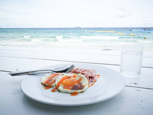 Aamiainen merinäköalalla tausta (paistettuja munia, kastiketta, munakas) Yksinkertaisesti aamiaista ruokaa puinen pöytä, kaunis trooppinen meri. Aamulla loma käsite. Jäähdytys matka käsite. - Valokuva, kuva