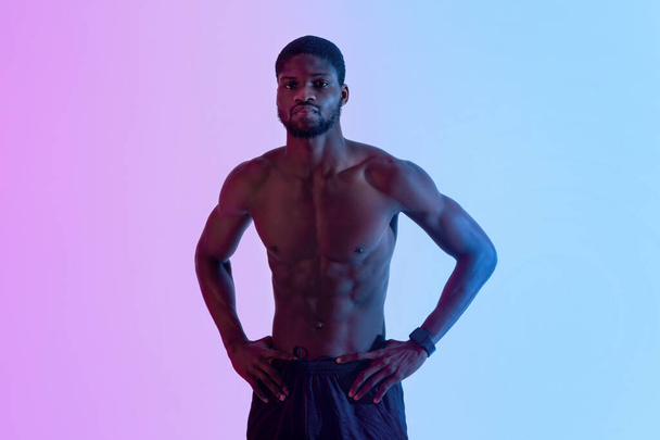 Muscular jovem homem negro com tronco nu posando com as mãos sobre os quadris em luz de néon - Foto, Imagem