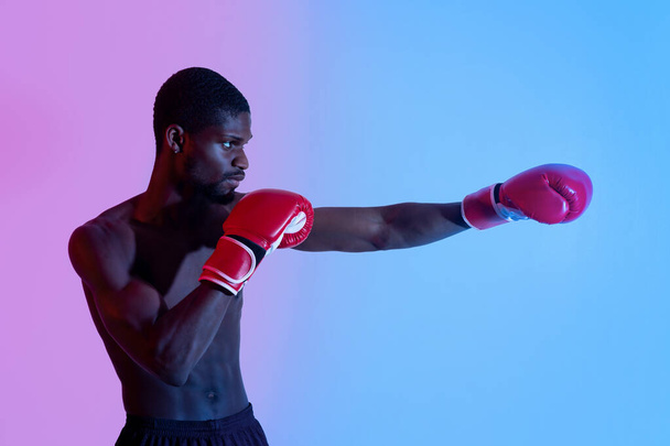 Zijaanzicht van professionele zwarte bokser in handschoenen gooien punch in neon verlichting - Foto, afbeelding