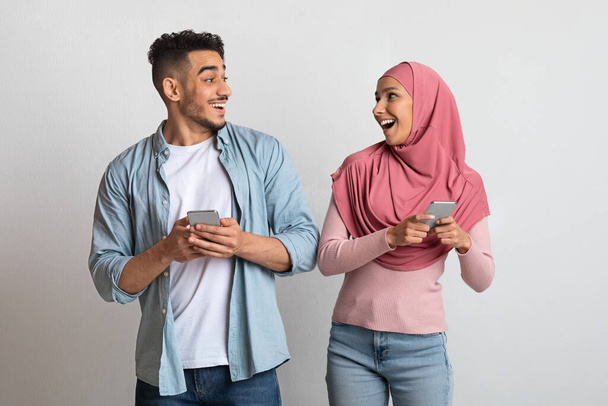 Online Offer. Excited islamic couple holding smartphones and looking at each other - Foto, Imagem