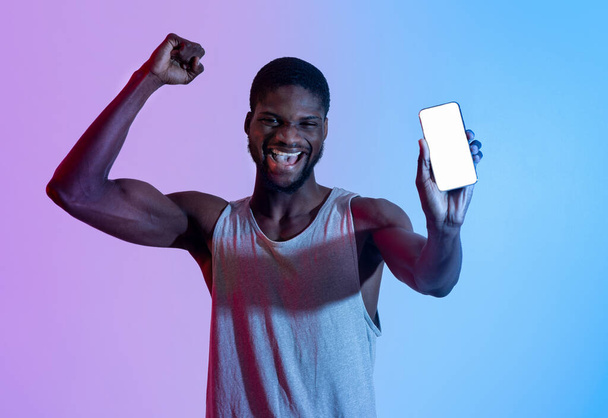 Aufgeregter schwarzer Athlet zeigt Handy mit leerem Bildschirm, macht JA-Geste im Neonlicht, Design-Attrappe - Foto, Bild