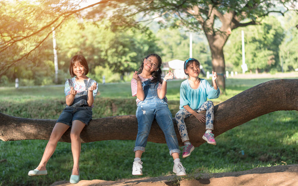 公園の幸せな女の子の子供たちとの子供の友情の概念は、学校の時間帯に友人と一緒に楽しい思い出と学生生活の瞬間を楽しんで一緒に遊んで木陰に座って楽しんでいます - 写真・画像