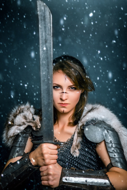 Portrait of a medieval woman warrior in chain mail and fur with a sword in her hands posing while standing in a snowstorm. Fantasy barbarian, viking. - Foto, Imagem