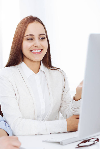 Νεαρή γυναίκα που κάθεται στο γραφείο με τον υπολογιστή στο ηλιόλουστο γραφείο. Φαίνεται σαν μαθήτρια ή επιχειρηματίας που επικοινωνεί με έναν απλό ντυμένο άντρα. - Φωτογραφία, εικόνα