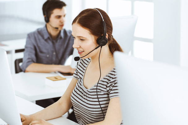 Casual dressed young woman using headset and computer while talking with customers online. Group of operators at work. Call center, business concept - Foto, Imagen