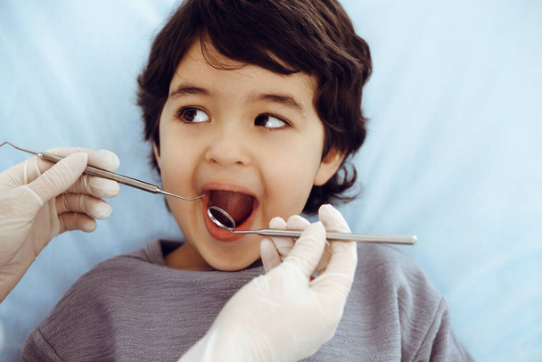 Netter arabischer Junge sitzt auf dem Zahnarztstuhl mit offenem Mund während der Oraluntersuchung beim Arzt. Zahnarztbesuche. Stomatologisches Konzept - Foto, Bild