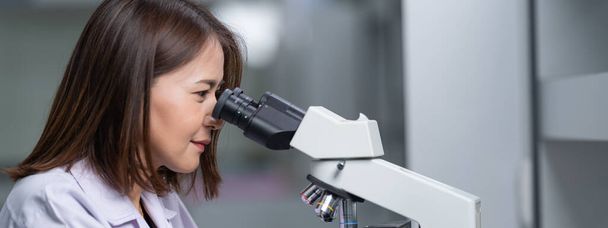研究室の顕微鏡を通して研究室のコートを着た若い科学者の女性が研究と実験を行う。研究室で働く科学者。教育ストックフォト - 写真・画像