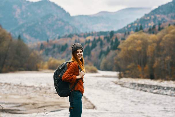 woman in a red sweater cap with a backpack on the river bank and mountains in the distance - Foto, Imagen
