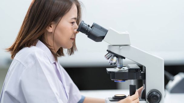 Młoda naukowiec kobieta w płaszczu laboratoryjnym patrząc przez mikroskop w laboratorium do badań i eksperymentów. Naukowiec pracujący w laboratorium. Zdjęcie edukacyjne - Zdjęcie, obraz