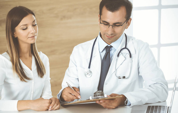 Doctor and patient discussing medical exam results while sitting at the desk in clinic. Male physician using clipboard for filling up medication history record of young woman. Data in medicine - Foto, Imagem