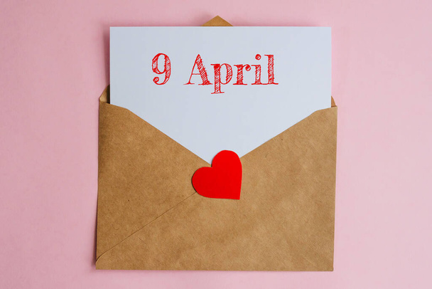 Kraft envelope with a white sheet of paper and a date 9 april, with red heart. Flat lay on pink background, romance and love concept - Фото, зображення