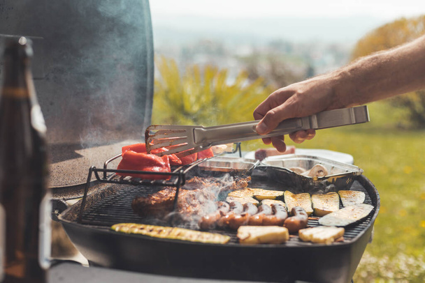 Close up of sausages, steak, cheese and vegetables on gas grill. Summer time, outdoors. - 写真・画像