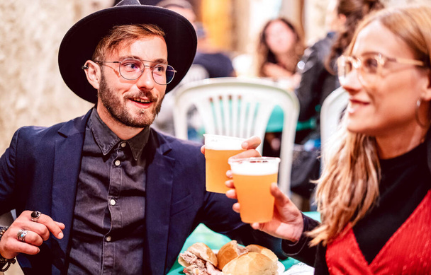 Young couple toasting beer glasses at street food festival - Beverage life style concept with friends having fun together on happy hour at brewery pub - Bright vivid filter with focus on guy with hat - Photo, Image