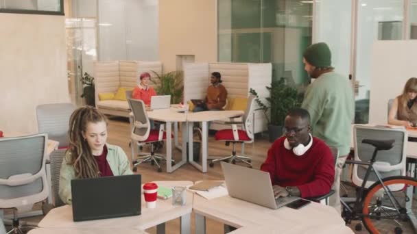 Inclinez-vous slow mo shot de divers jeunes entrepreneurs travaillant sur des ordinateurs portables dans un bureau décloisonné de start-up créative ou un espace de coworking. Homme barbu en bonnet chapeau parking son vélo dans le bureau - Séquence, vidéo