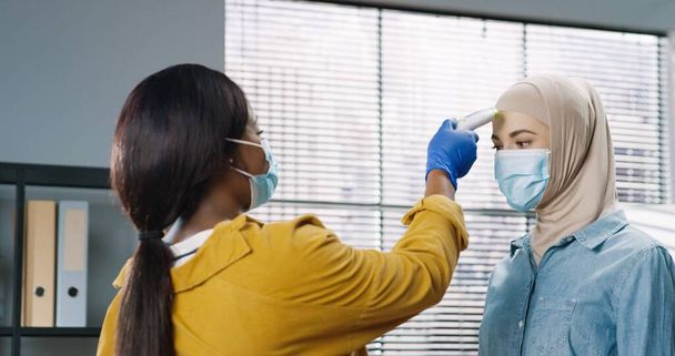 Posteriore della femmina afroamericana in maschera medica in piedi in ufficio a controllare la temperatura di misurazione della salute dei lavoratori utilizzando il termometro a infrarossi durante la pandemia di coronavirus. Concetto di quarantena ravvicinata - Foto, immagini