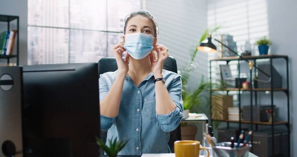 Portrait of young beautiful girl in blue shirt sitting in office at table and takes off protective medical mask smiling looking at camera. - Фото, зображення