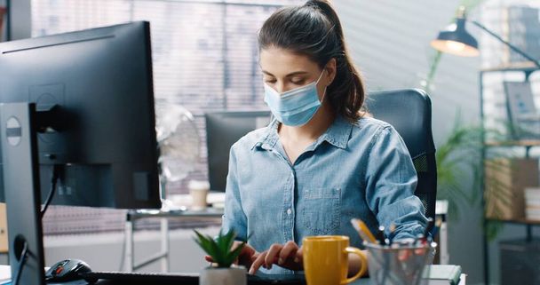 Close up portrait of beautiful young concentrated businesswoman in medical mask sitting at desk and typing on computer searching internet at office in cabinet. Corporate work, job concept - Фото, зображення