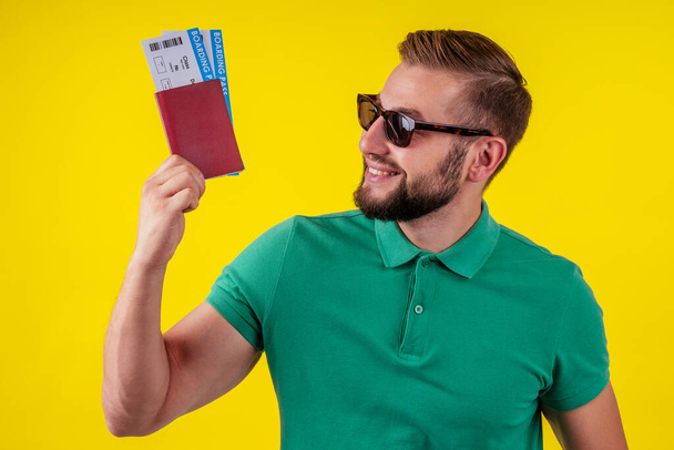 ortrait of a happy young man in sunglasses and green t-shirt holding passport isolated over yellow background - 写真・画像