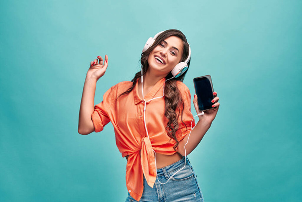 Beautiful young woman in an orange shirt dances laughs and shows the phone to the camera and listens to music in white headphones on a blue background. Lifestyle and good mood! - Photo, image