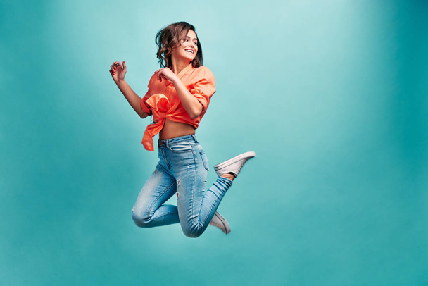 Young beautiful woman who is dressed in an orange shirt and blue jeans bouncing on a blue background with happiness and looking away and laughing. Emotion of admiration and freedom. - Foto, afbeelding