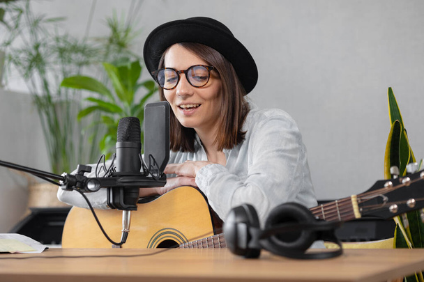 podcast, music audio content creation. beautiful European woman podcaster in a hat with a guitar or ukulele, radio host recording podcast or content, song and music in a recording studio - Valokuva, kuva
