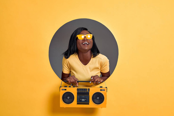 Image of beautiful young woman in yellow sunglasses laughing and holding vintage boombox isolated over circle hole in yellow background. Copy space. - 写真・画像