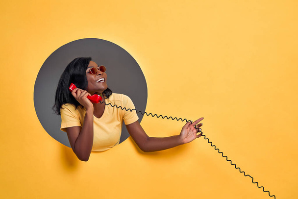 Portrait of a smiling happy afro american woman in sunglasses talking on retro telephone in a round hole circle in yellow background. Copy space. - Zdjęcie, obraz