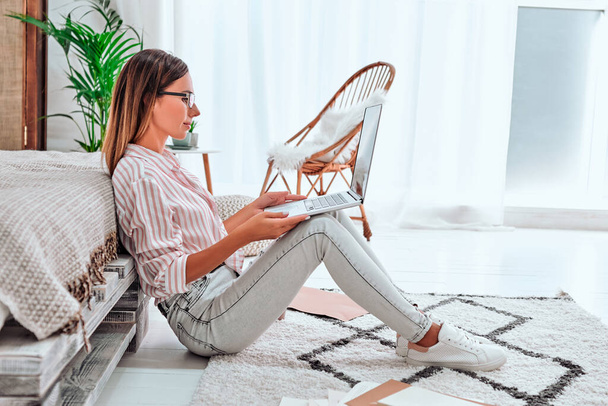 Putting fresh content into her blog. Thoughtful young woman in eyewear working using computer while flooring at home. Side view - Foto, afbeelding