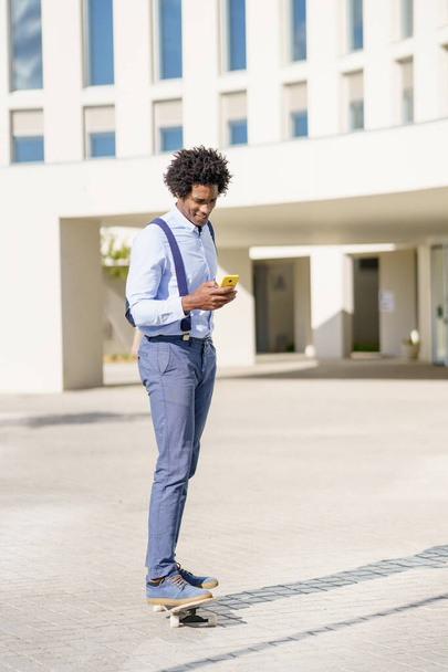 Μαύρος επιχειρηματίας σε ένα skateboard κοιτάζοντας smartphone του σε εξωτερικούς χώρους. - Φωτογραφία, εικόνα