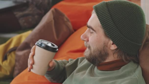 Κοντινό πλάνο του χαρούμενου γενειοφόρου άνδρα σε καπέλο beanie ανακλινόμενα στην καρέκλα τσάντα φασολιών και πίνοντας καφέ από-go κύπελλο - Πλάνα, βίντεο