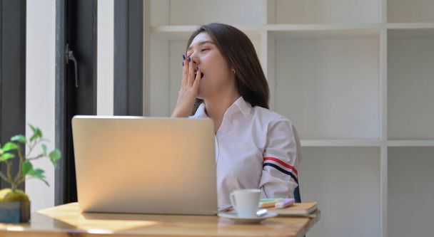 Young woman with a laptop and coffee in the office. She covered mouth with hand and yawned while resting. - Фото, изображение