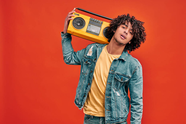 Waist up portrait of young handsome stylish man with bright beat box on his shoulder on isolated orange. Eastern boy in jeans clothes with retro music player - Foto, Imagen