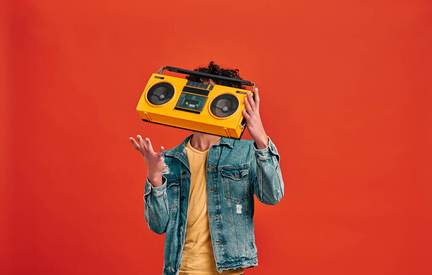 Studio shot of man catching or throwing retro music player, which hiding his face. Isolated on red background - Фото, изображение