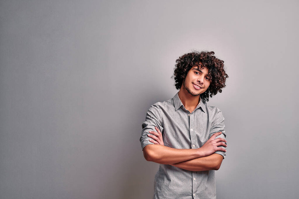 Smiling arabian boy in a striped casual shirt with curly hairsyle standing against gray background. Isolated image of smiling eastern young man with crossed hands - Foto, Bild