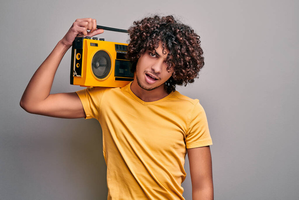Young handsome eastern student with retro tape stereo player on his shoulder standing on gray isolated background - Foto, afbeelding