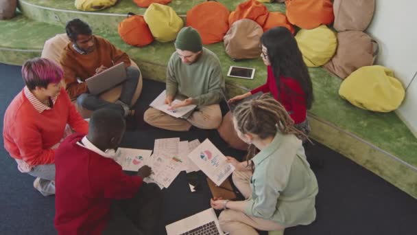 Hoge invalshoek van multi-etnische groep jonge ondernemers die op de vloer van de loungeruimte in het startkantoor zitten en financiële documenten bespreken - Video