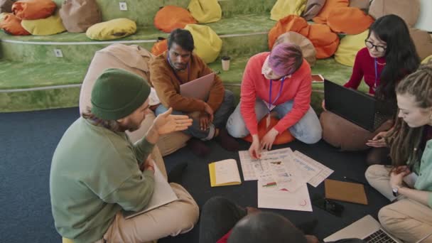 スタートアップオフィスの快適なラウンジエリアの床に座っている若い男性と女性の多様なグループの高い角度で撮影されたPANスローモやプロジェクトに取り組んでいる間に文書を議論 - 映像、動画