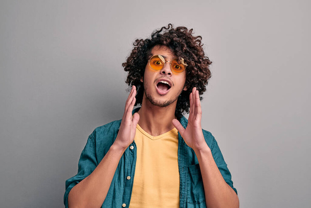 Portrait of surprised young eastern man screaming with hands cupping his mouth on isolated gray background. Shouting stylish casual student in eyeglasses - Foto, Bild