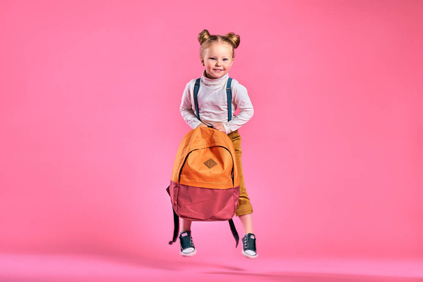 Funny little girl with big backpack jumping and having fun against pink wall. School concept. Back to School. School's out for summer. Celebrating the end of another successful school year. Copy space - Zdjęcie, obraz