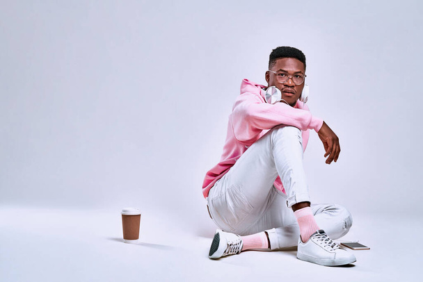 Lifestyle concept. Young African American man wearing a pink hoodie, white pants and pink socks posing in the studio with headphones, a telephone and coffee. Concepts of style, technology, gadgets. - Photo, image