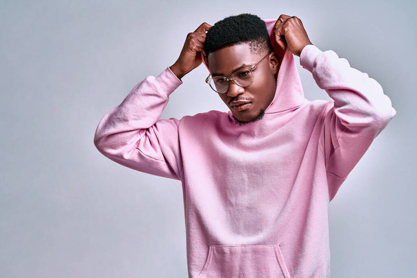 Close up of young african american man in pink streetwear hoodie with hood on head posing isolated on grey background in studio. People lifestyle concept. Copy space. Looking aside down - Valokuva, kuva