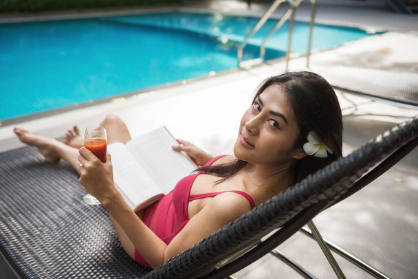 Portré ázsiai gyönyörű fiatal nő piros fürdőruhában olvasni könyvet és inni gyümölcslevet az ágy szék az uszodában. Szexi lány nyaralás készítő a nyári vakáció a luxus hotel resort. - Fotó, kép