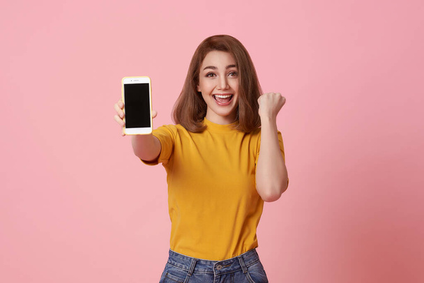 Ευτυχισμένη νεαρή γυναίκα δείχνει σε κενή οθόνη κινητό τηλέφωνο και χέρι χειρονομία επιτυχία απομονώνονται σε ροζ φόντο. - Φωτογραφία, εικόνα