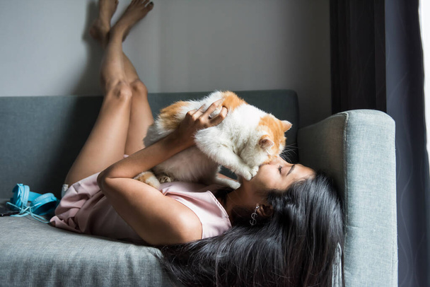 Ασιάτισσα νεαρή γυναίκα φιλάει και αγκαλιάζει την κίτρινη εξωτική κοντή γάτα της στον καναπέ του σαλονιού. κορίτσι μαζί με το κατοικίδιο ζώο της. Ο καλύτερος φίλος.. - Φωτογραφία, εικόνα