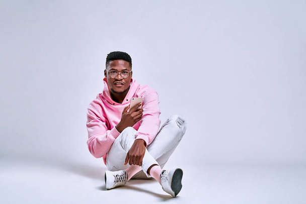 Studio fashion portrait of attractive young man in pink hoodie and blue jeans. Guy sits on floor over grey background. Copy space - Foto, Imagem